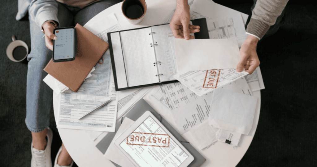 Two people sitting at a table covered with financial documents, a tablet displaying the word PAST DUE, envelopes stamped PAST DUE and LAST DUE, a notebook, coffee cup, and a smartphone with a calculator app open.