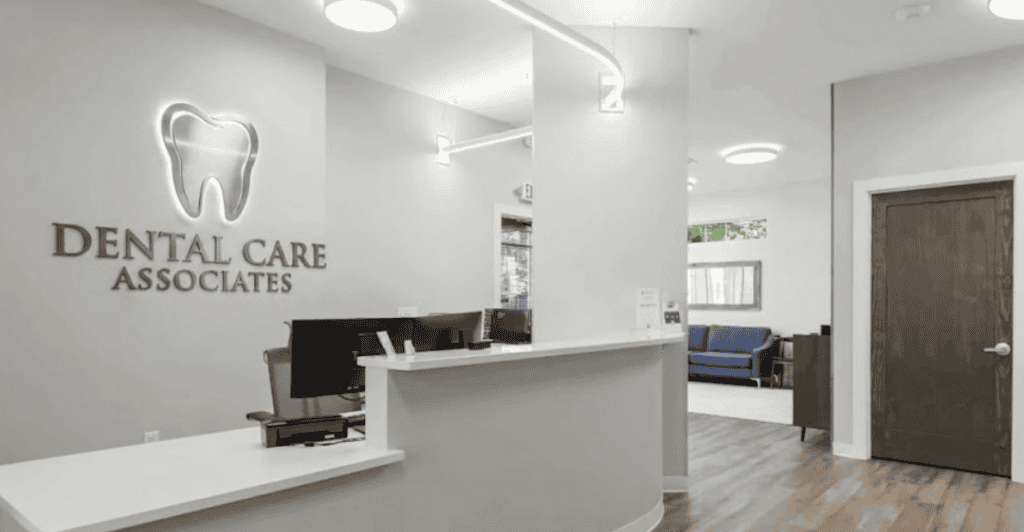 Modern dental office reception area with a white desk, computer monitors, a wall sign reading Dental Care Associates with a tooth icon, wooden floors, and a waiting area with blue chairs in the background.