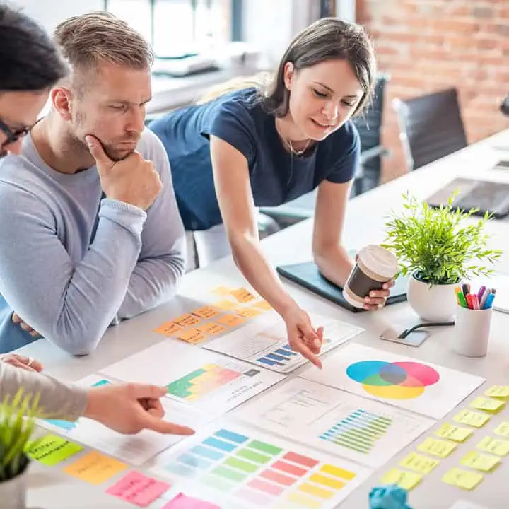 Four colleagues work together at a desk with colorful charts, graphs, sticky notes, and a plant. One woman points at a chart while holding coffee, and the group discusses data and plans.