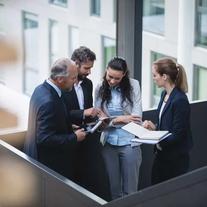 Four business professionals stand in a modern office hallway, gathered closely while looking at a digital tablet and taking notes, engaged in a collaborative discussion.