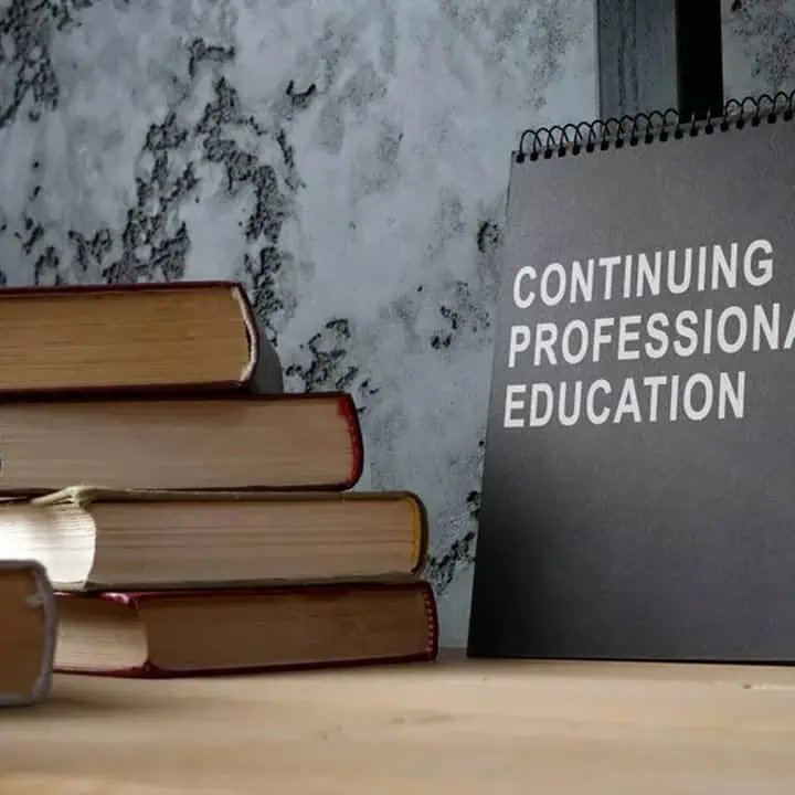 A stack of books sits on a wooden surface next to a black sign with white text that reads CONTINUING PROFESSIONAL EDUCATION, against a textured gray wall.
