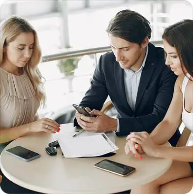 Three people sit at a round table with documents, pens, and smartphones. A man in a suit looks at his phone while two women, one in a blouse and one in a dress, listen and look at the papers.