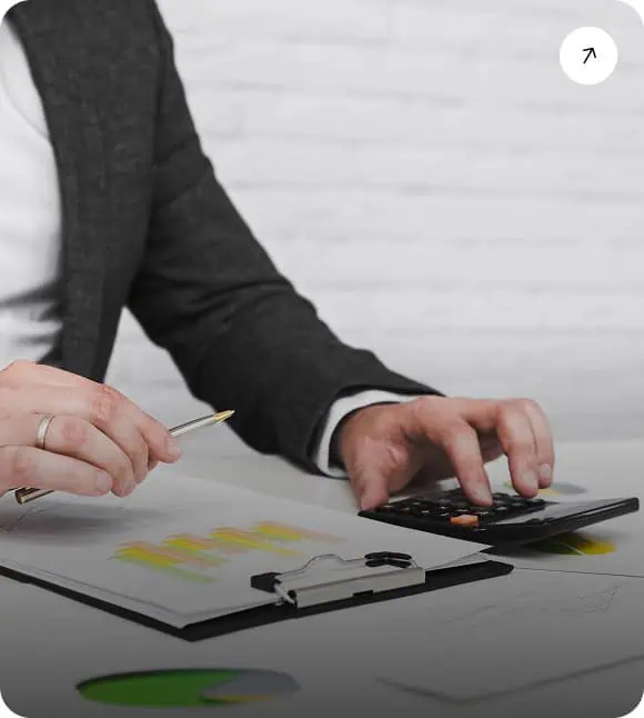 A person in a suit uses a calculator with one hand and holds a pen in the other, reviewing bar graphs and charts on a clipboard at a desk.