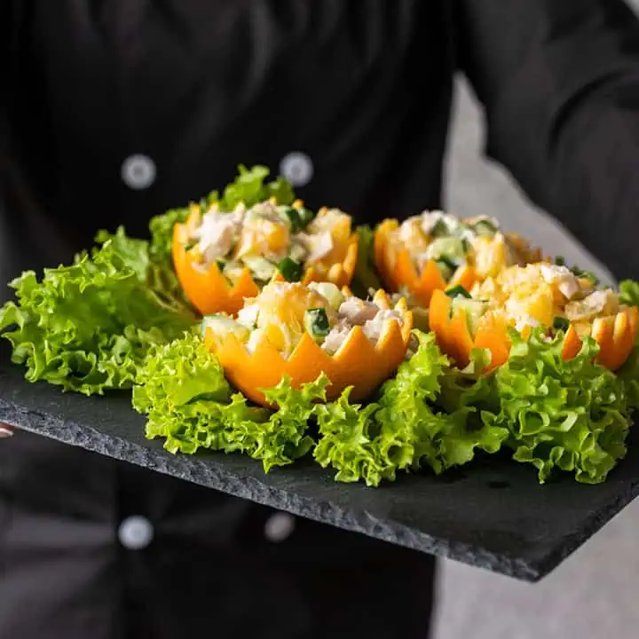 A person in a black chefs coat holds a slate tray with salad served in orange peel cups, garnished with chopped greens and surrounded by fresh lettuce leaves.