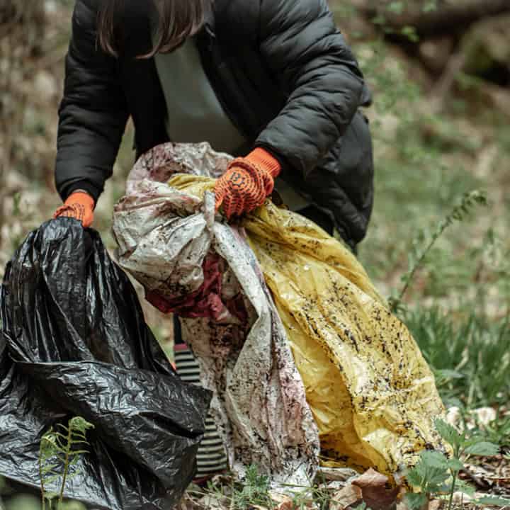 A person wearing an insulated jacket and orange gloves picks up large dirty plastic bags from the ground in a grassy outdoor area, cleaning up litter.
