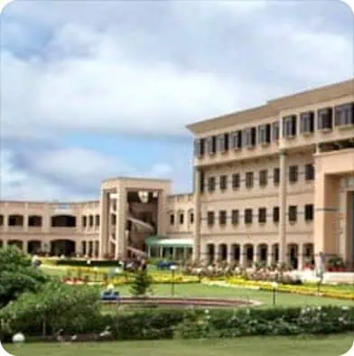 A large, modern beige building with multiple floors and rectangular windows, surrounded by green lawns, pathways, and landscaped gardens under a partly cloudy sky.