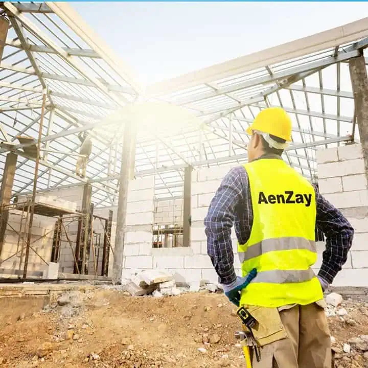 A construction worker in a yellow safety vest and hard hat, labeled AenZay, stands with hands on hips, observing a building under construction with exposed metal framework and sunlight in the background.