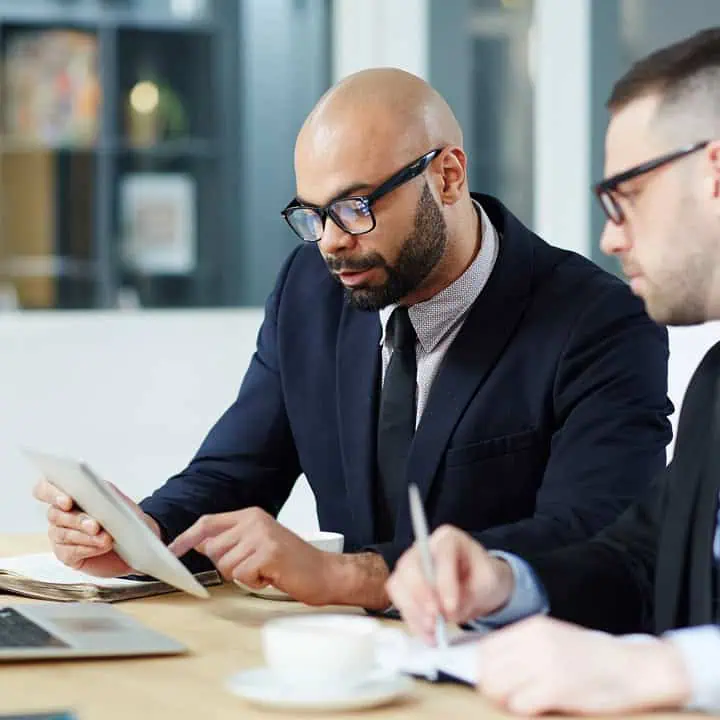 Two men in business suits sit at a table in an office, one looking at a tablet and the other writing notes on paper. A laptop and a cup are on the table in front of them.
