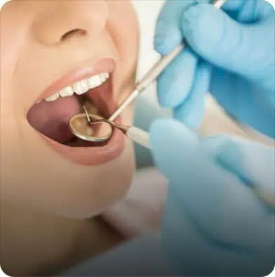 A dentist wearing blue gloves examines a patient’s open mouth with a dental mirror and instrument.