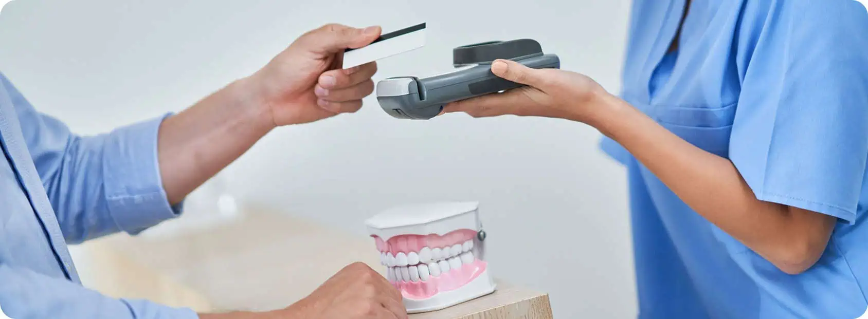 A person hands a credit card to a healthcare worker holding a card reader, with a dental model displaying teeth on the counter—illustrating how dental debt collection services can streamline payment processes in dental practices.