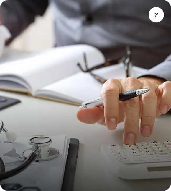 A person holding a pen uses a calculator on a desk with an open notebook, eyeglasses, and a stethoscope, suggesting work involving financial or medical calculations.