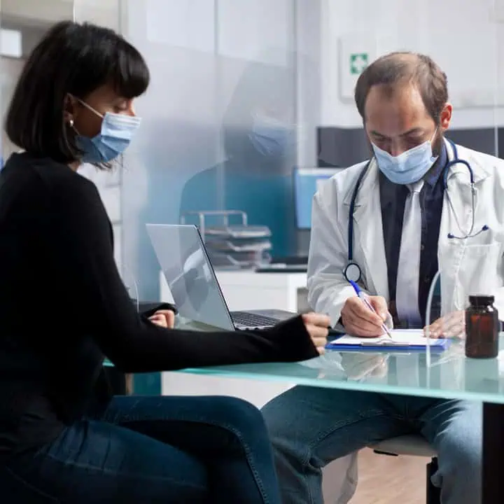 A woman wearing a mask sits at a glass desk across from a masked doctor with a stethoscope, who is writing on a clipboard in a modern medical office.