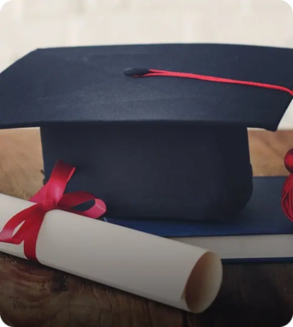 A graduation cap with a red tassel and a rolled-up diploma tied with a red ribbon rest on a wooden surface.