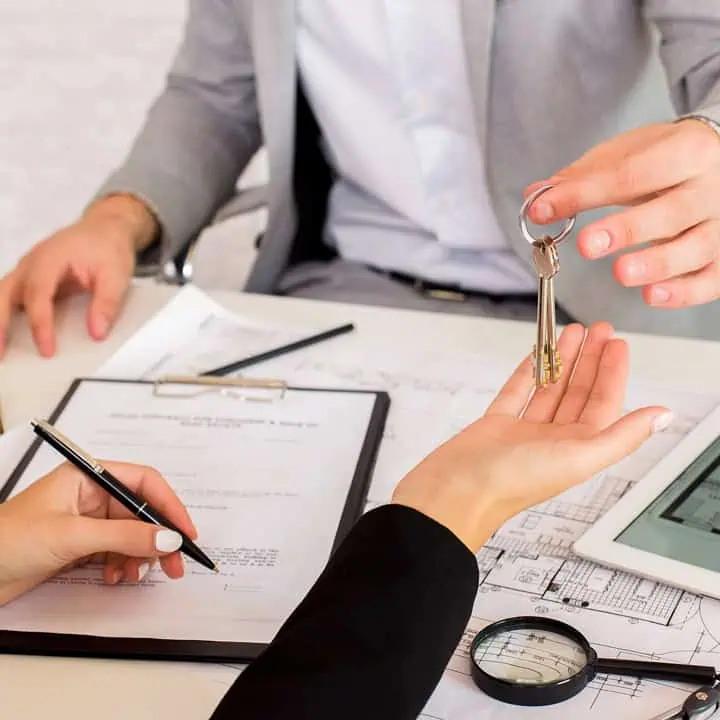 A person hands a set of keys to another person over a desk with documents, a clipboard, a pen, and a tablet, suggesting a real estate or property transaction.