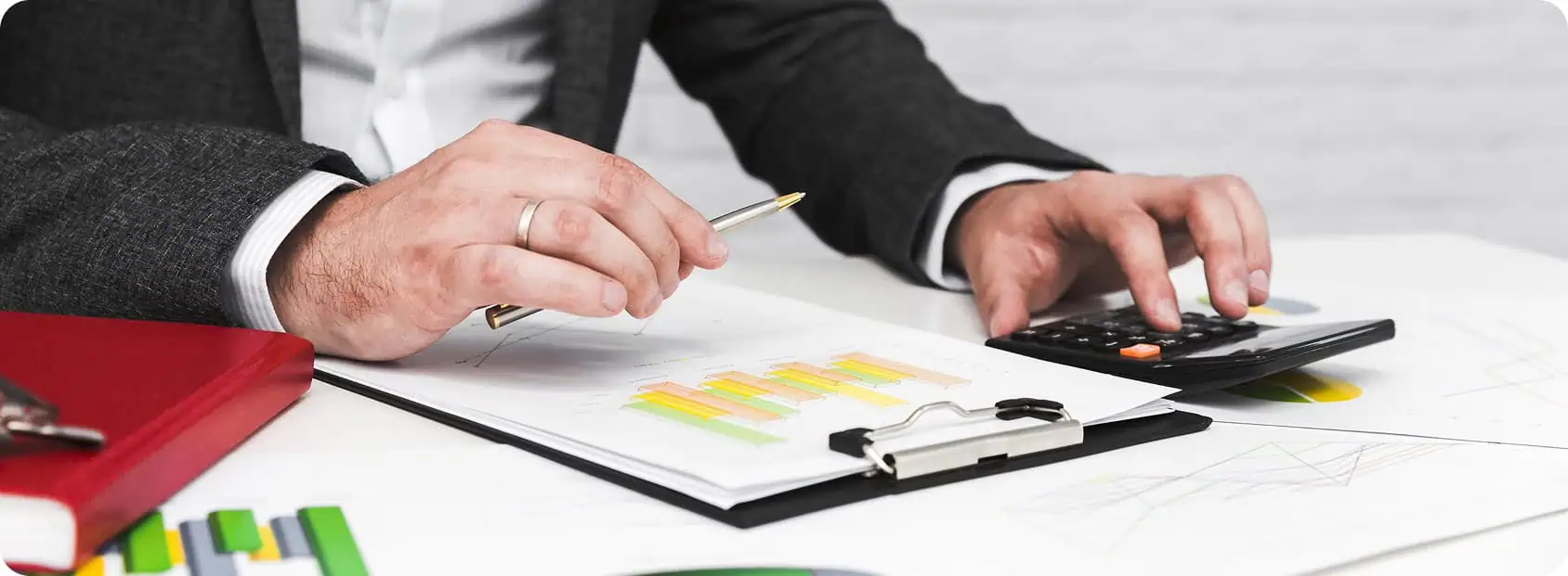 A person in a suit is working at a desk with financial charts, reviewing professional service debt collection documents, holding a pen over bar graphs and using a calculator. Other papers and a red notebook are also on the desk.