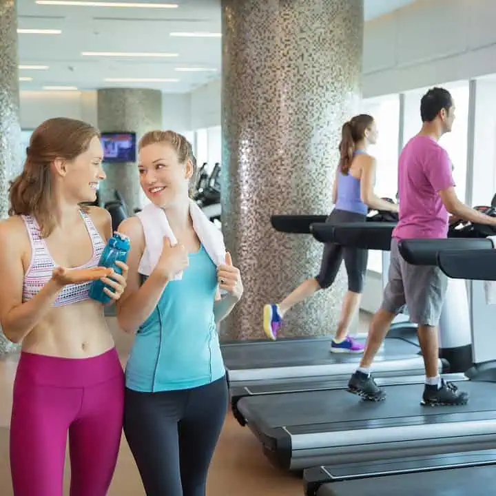Two women in workout clothes stand by treadmills, smiling and talking. One holds a water bottle, the other has a towel around her neck. In the background, a man and another woman are running on treadmills in a bright gym.
