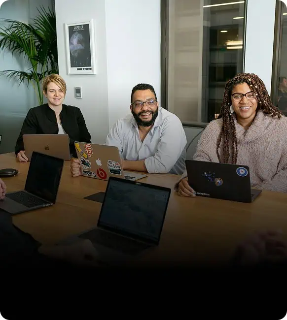 Three people sit at a conference table with laptops, smiling and engaged in a meeting. A plant and framed picture are in the background, and daylight streams through the window.