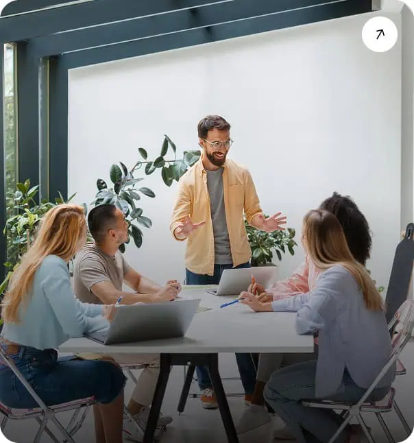 A man stands and speaks enthusiastically to four colleagues seated around a table with laptops and notebooks in a bright, modern office with large windows and green plants.