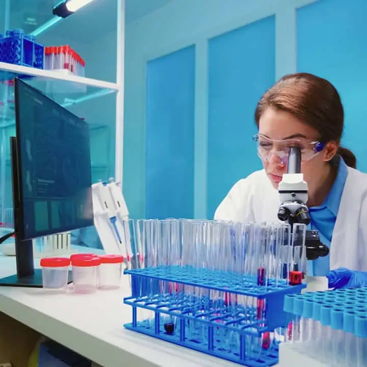 A scientist wearing safety goggles examines samples through a microscope in a laboratory, surrounded by racks of test tubes and sample containers, with a computer monitor and lab equipment nearby.