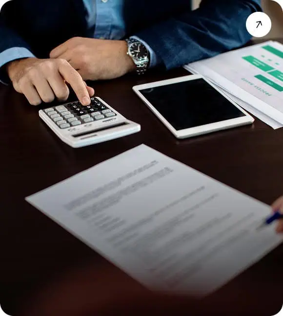 A person in business attire uses a calculator at a desk with documents, a tablet, and charts, suggesting financial or business planning activities.