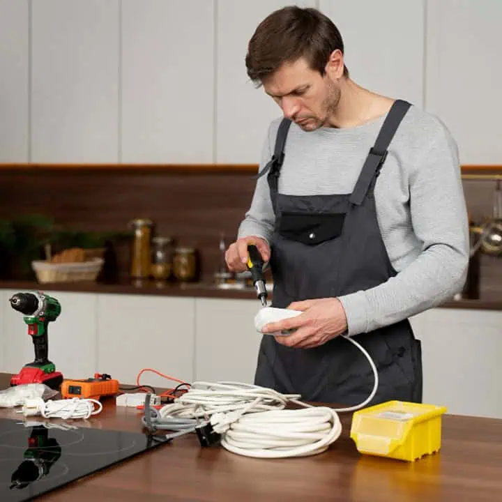 A man in overalls uses pliers to work on an electrical plug at a kitchen counter, surrounded by tools, cords, and a yellow toolbox.