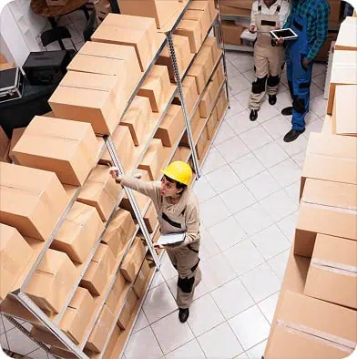 A worker wearing a yellow hard hat and beige uniform inspects boxes on a high shelf in a warehouse, holding a clipboard. More boxes and two other workers are visible in the background.
