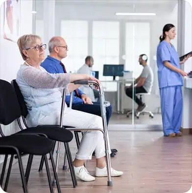 Older woman with a walker sits in a waiting room with two other people. Medical staff in scrubs work at a computer in the background behind glass walls. The setting appears to be a healthcare facility.