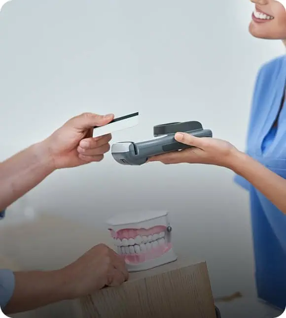 A person in scrubs holds a card payment terminal while another person pays with a credit card; a dental model with teeth is visible on the table between them.