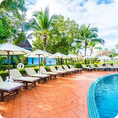 A row of empty lounge chairs with white umbrellas lines the edge of a curved outdoor pool, surrounded by lush green trees and palm trees under a partly cloudy sky.