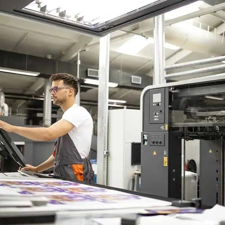 A man wearing glasses and work overalls operates a large printing machine in a modern industrial print shop, with colorful printed sheets visible on the counter beside him.