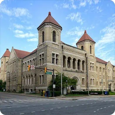 Historic stone building with two red-roofed towers, arched windows, and a corner entrance, situated at a street intersection with traffic lights under a partly cloudy blue sky.