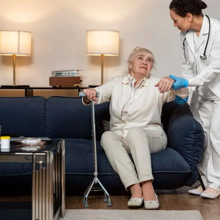 An elderly woman with a cane sits on a couch, looking up at a healthcare worker in a white coat and gloves who is helping her stand. There are lamps, a table, and medical supplies nearby.
