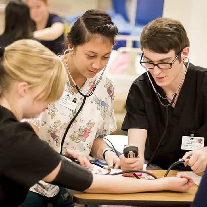 Two medical students wearing scrubs and stethoscopes check a patient’s blood pressure in a clinical setting, focusing on the procedure and recording results, while another person sits with her arm extended.