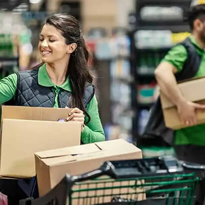 A woman in a green shirt and black vest is smiling while stocking shelves with cardboard boxes in a grocery store. A man in similar attire carries boxes in the background. A shopping cart is in the foreground.