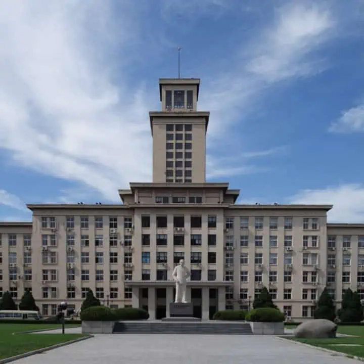 A large, beige building with symmetrical windows and a central tower stands under a blue sky with scattered clouds. In front of the building is a statue of a person on a pedestal, surrounded by greenery.