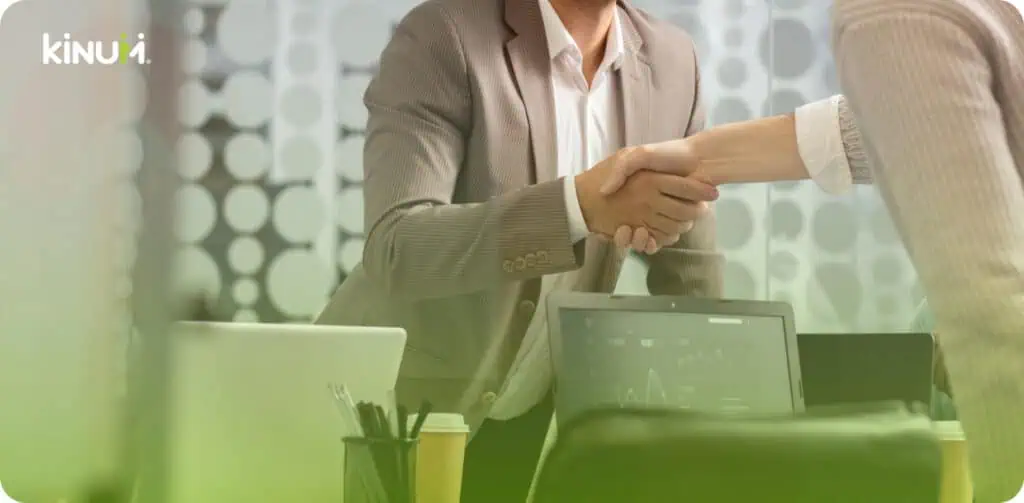 Two people in business attire shake hands across a desk with laptops, papers, and coffee cups. The background features frosted glass with circular patterns. The kinui logo is visible in the top left corner.