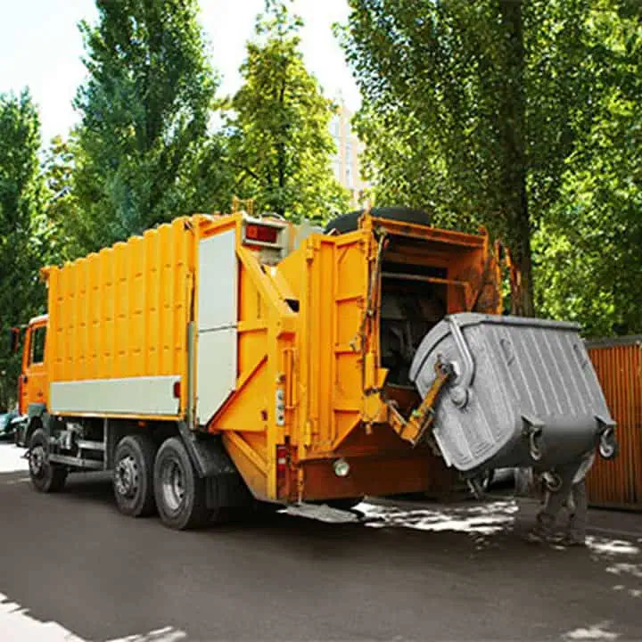 A large yellow garbage truck lifts and empties a metal trash bin on a tree-lined street in daylight.