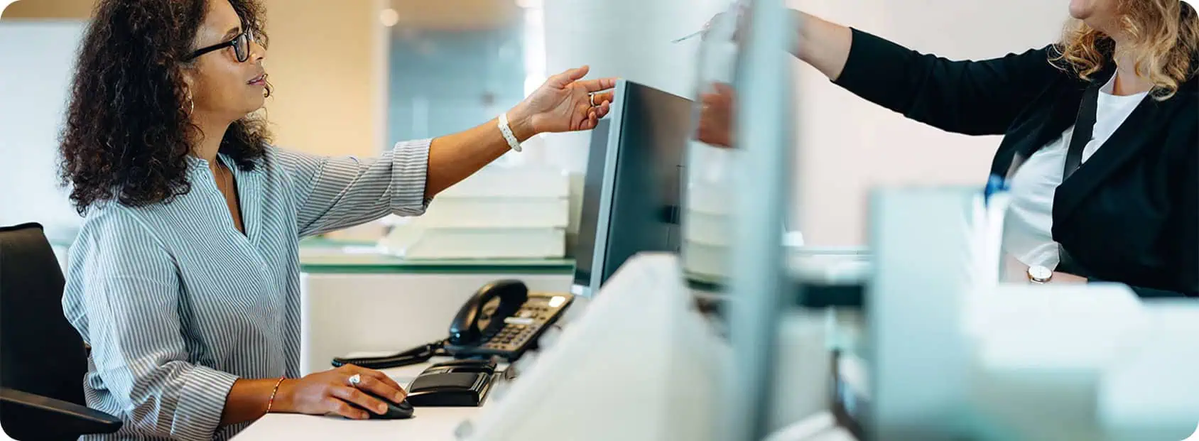 Two women interact at an office front desk; one is seated at a computer, reaching for a document, while the other stands, handing over public sector collection recovery paperwork across a glass divider.