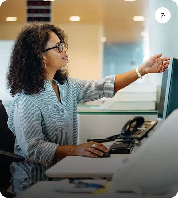 A woman wearing glasses sits at a desk, using a computer while reaching out to examine a sheet of paper on a wall or board in a modern office setting.